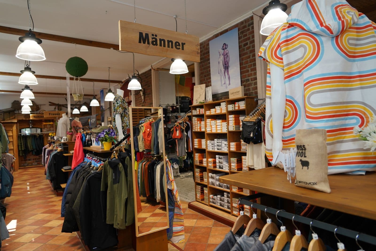 Innenraum eines Modegeschäfts mit Kleidungsständern, Regalen und buntem Dekorationsstoff in Wellenmuster, Beschilderung 'Männer'