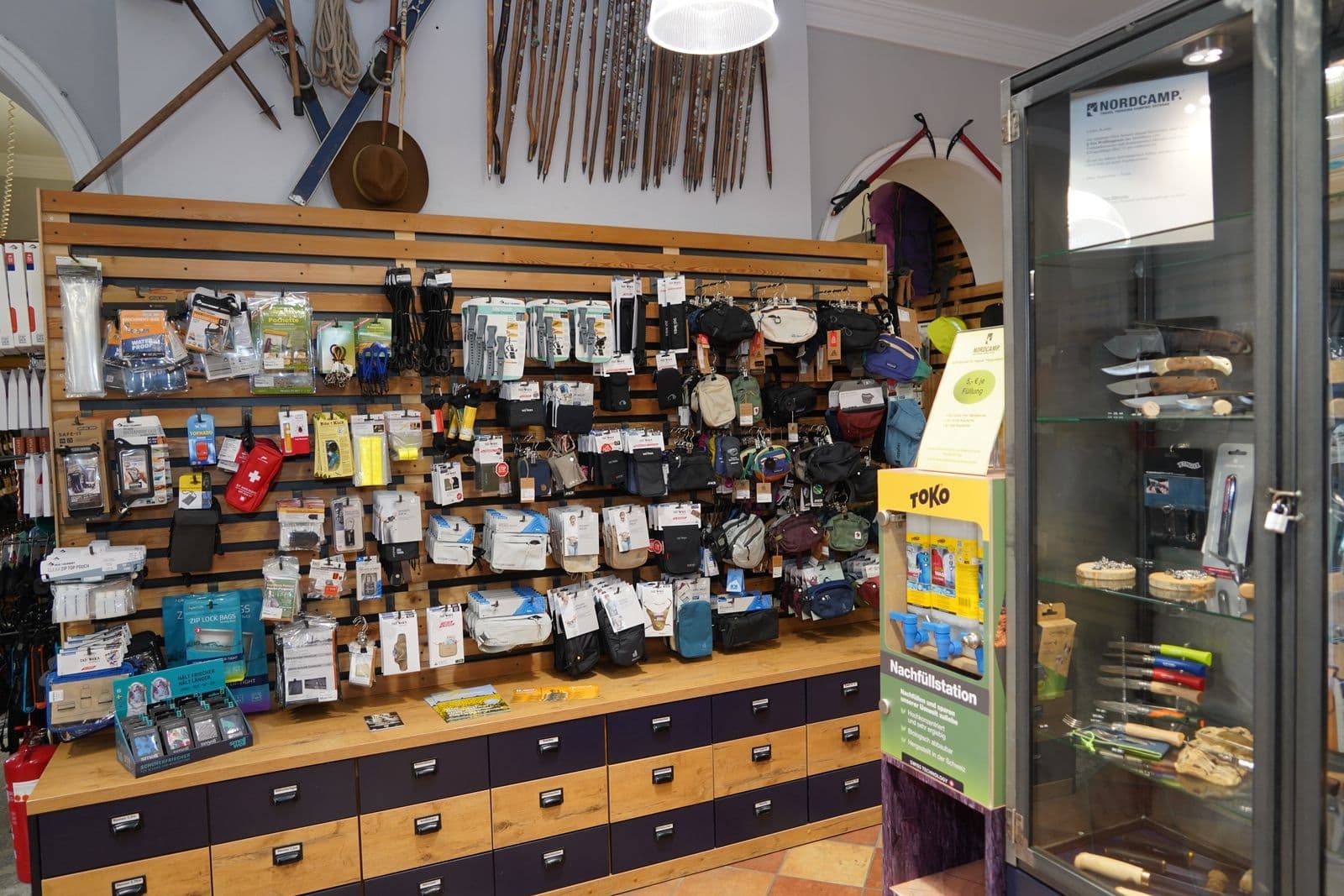 Ladentheke eines Outdoor- und Campingfachgeschäfts mit umfangreicher Ausstellung von Ausrüstung, Rucksäcken, Werkzeugen und Zubehör an Lochplatten und auf Holztheke.