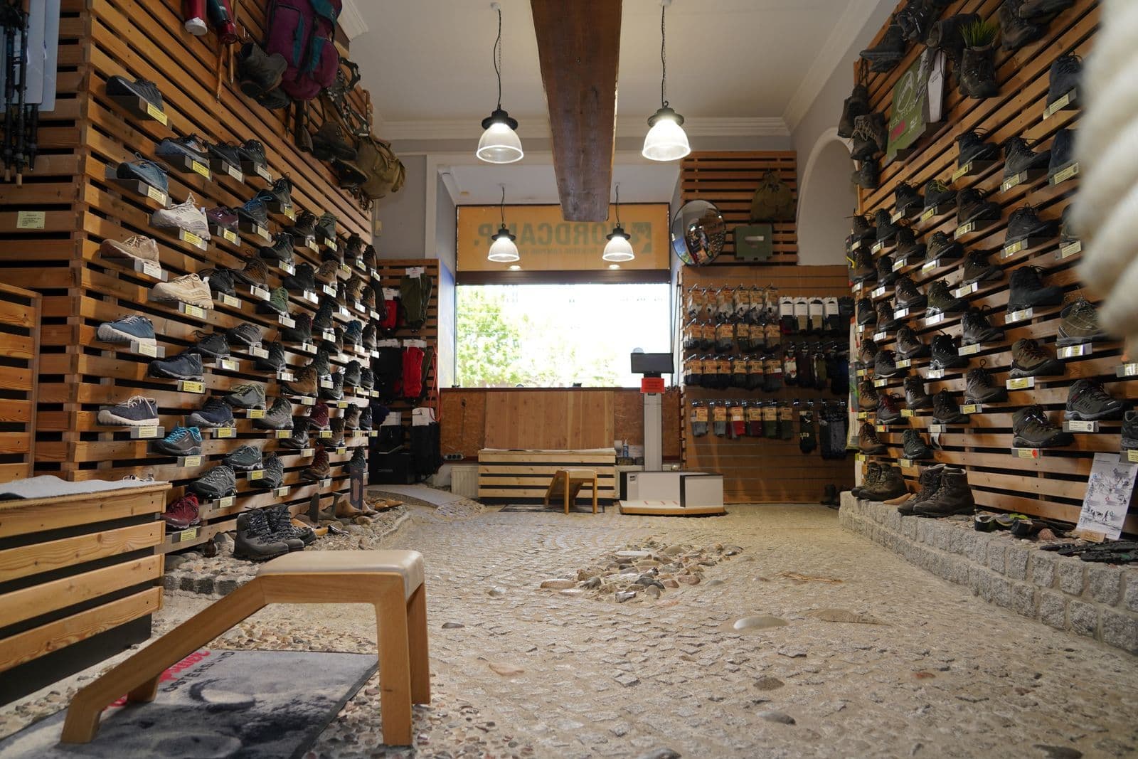 Innenraum eines Outdoor-Fachgeschäfts mit Schuhen und Ausrüstung auf Holzpalettenregalen, Sandböden und Industriebeleuchtung.